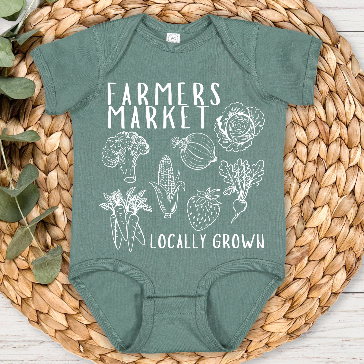 Green baby onesie with 'Farmers Market Locally Grown' text and vegetable illustrations on a woven mat.